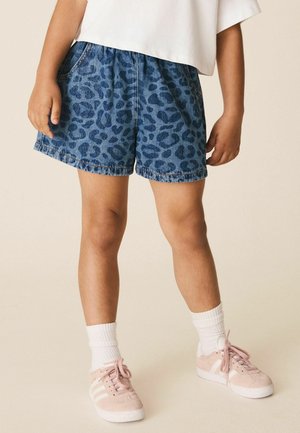 Kind trägt blaue Denim-Shorts mit Leopardenmuster, weiße Knöchelsocken, pinke Sneakers und ein weißes Top, das auf Taillenhöhe geschnitten ist.