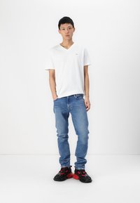 T-shirt blanc à manches courtes en V, jeans bleu clair, baskets épaisses noires et rouges. Le mannequin se tient devant un fond blanc uni.
