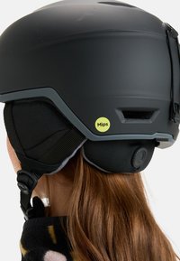 Schwarzer Skihelm mit matter Oberfläche, ausgestattet mit einem MIPS-Label, verstellbaren Belüftungsöffnungen und Ohrpolsterung, gesichert mit einem Schnallenriemen.