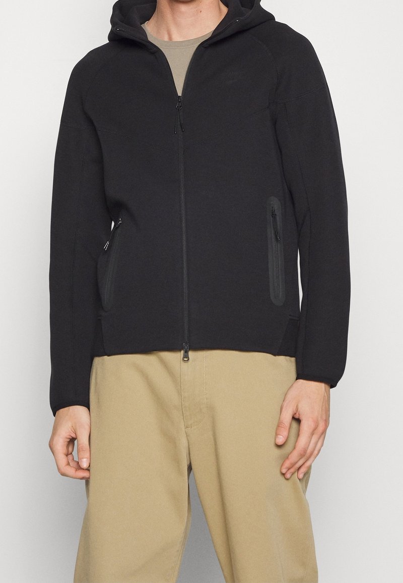 Svart zip-up hoodie i mjukt material med huva, sidofickor med dragkedja och en åtsittande design, parat med beiga byxor.