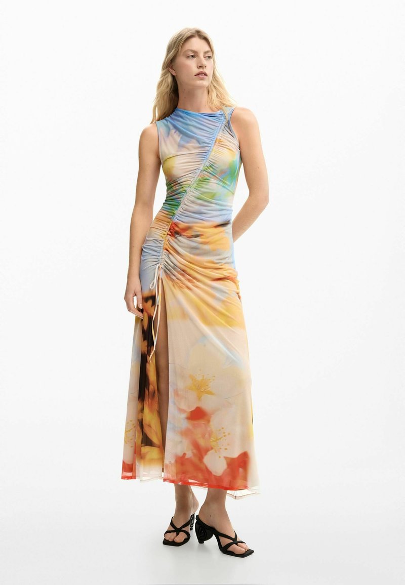 Frau trägt ein ärmelloses, gerafftes Maxikleid mit mehrfarbigem Blumenmuster und einem seitlichen Schlitz, kombiniert mit schwarzen Riemchen-Sandaletten mit Absatz.