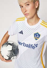 Baltas futbola kreklis ar ziliem un dzelteniem akcentiem, kuram ir diagonālas svītras un logotipi. Spēlētājs tur melno un baltu rakstu futbola bumbu.
