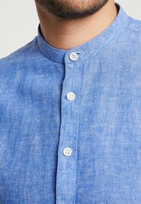 Lichtblauw linnen overhemd met een opstaande kraag, voorzien van vijf witte knopen aan de voorkant. De stof heeft een gestructureerde, licht rommelige uitstraling.
