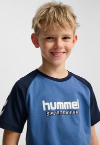Blå och marinblå T-shirt med raglanärmar. Har logotypen "hummel SPORTSWEAR" i vitt. Mjukt bomullsmaterial, rund halsringning.