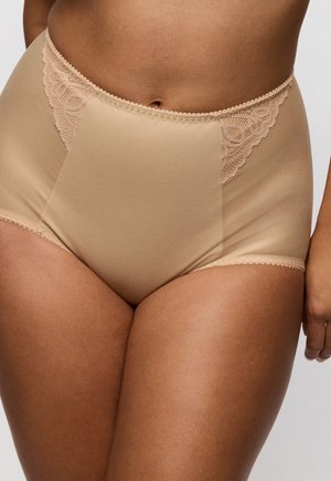 Vrouw die beige hoog uitgesneden ondergoed met kanten accenten op de heupen draagt, staand tegen een effen achtergrond.