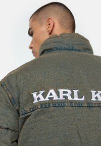 Sötétzöld farmerdzseki magas gallérral és a háton nagy, fehér hímzett "KARL K" logóval. Texturált anyag, laza szabás.