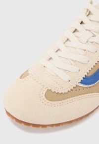 Beige semsket sneaker med blå logo-detalj, beige detaljer og flate hvite lisser. Glatt tekstur med rund tå og gummisåle.