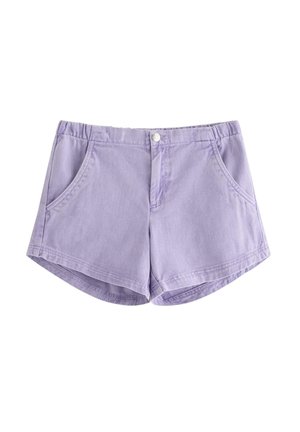 Ung pige står, iført en hvid kortærmet top med små blå mønstre, lavendelfarvede shorts, hvide sokker og hvide sneakers.