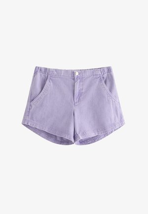 Młoda dziewczyna stoi, ubrana w biały krótki top z małymi niebieskimi wzorami, lawendowe szorty, białe skarpetki i białe trampki.