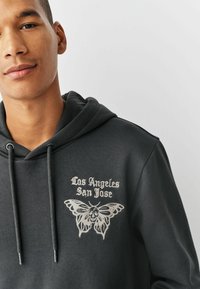 Donkergrijze hoodie met een trekkoord, voorzien van een grafisch ontwerp van een schedel en een vlinder, samen met de tekst "Los Angeles San Jose" in witte borduursel.