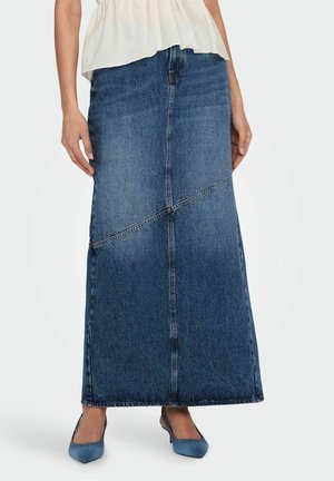 Mujer con una falda larga de denim azul con detalle de costura diagonal, top blanco y zapatos slingback azules con punta puntiaguda sobre un fondo liso.