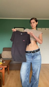 Fekete, rövid ujjú póló, melynek mellkasán egy apró piros logó található, zöld fal előtt kiállítva. A modellen világos mosású, laza farmernadrág van.