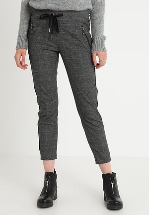 Personne portant un pantalon cintré à carreaux gris avec une taille à cordon noir, des poches latérales zippées noires, un pull gris et des bottines noires à fermeture éclair devant.