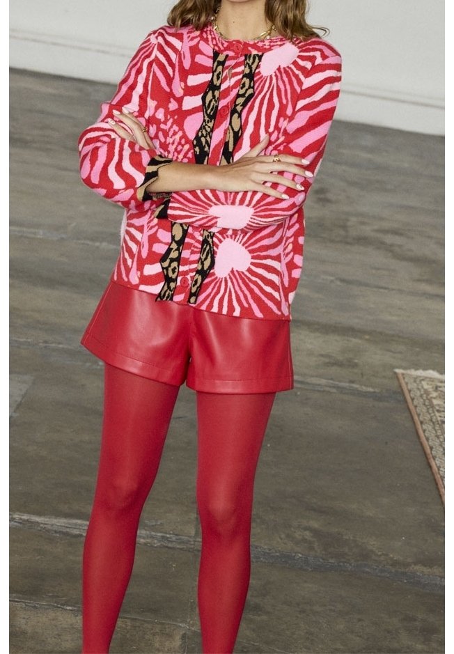Suéter de patrón rojo con remolinos rosas y blancos, combinado con pantalones cortos de cuero rojo y leggings rojos. Destacan los detalles en negro y dorado.