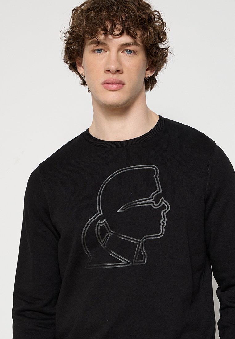 Pull noir avec un contour en relief texturé, de la forme d'un profil, sur le devant. Il présente un col rond et des manches longues.