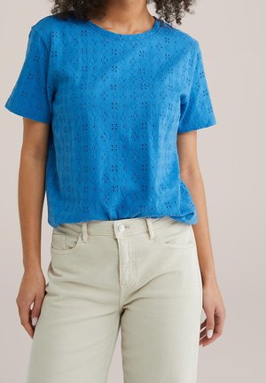 Blouse bleue à manches courtes avec des broderies œillet et des motifs floraux, portée avec un pantalon taille haute blanc cassé sur un fond neutre.