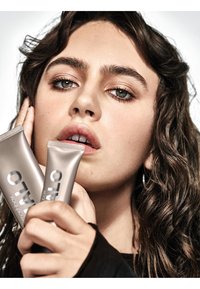 Dos tubos plateados del producto "SMASHBOX" sostenidos cerca de la cara, con un fondo desenfocado suave. El cabello es ondulado y oscuro, con un maquillaje natural.