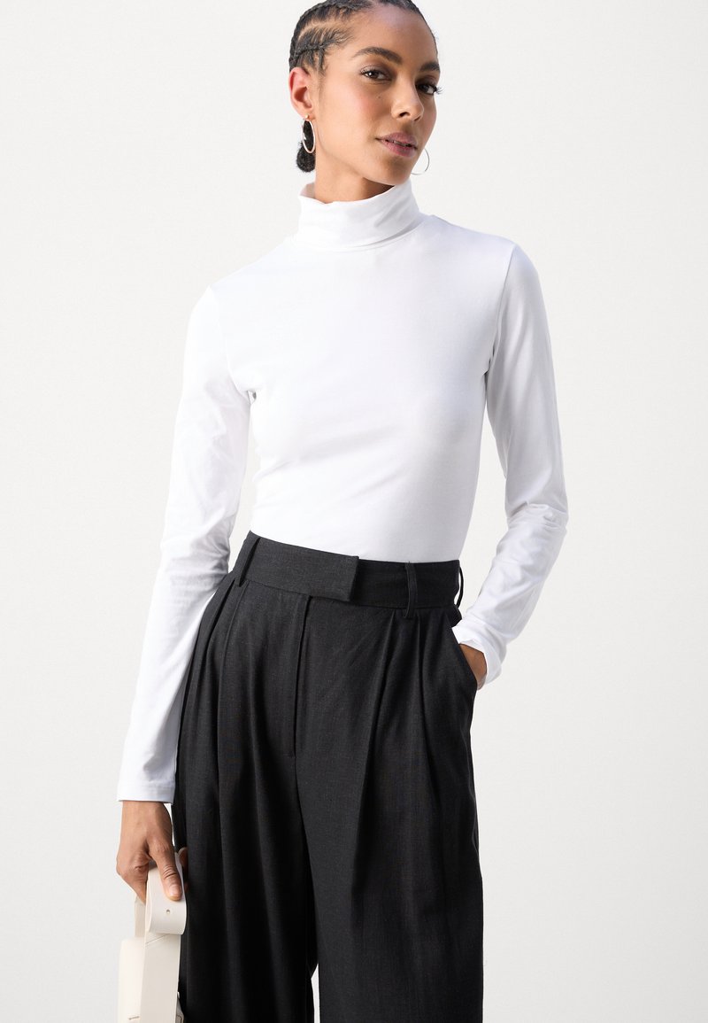 Maglione bianco a maniche lunghe con collo alto abbinato a pantaloni neri a pieghe a vita alta. L'outfit è completato da una borsa bianco sporco e orecchini a cerchio.