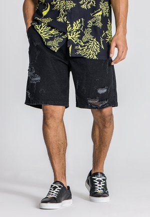 Black denim shorts with distressed detailing, featuring frayed edges and rips. Paired with a patterned shirt and black sneakers.
