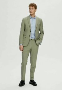 Fato verde claro com calças ajustadas e um casaco de botão único. Combinado com uma camisa azul clara e sapatos pretos. Textura suave e design sob medida.