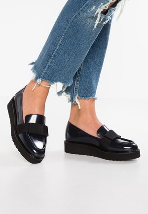 Mocassins compensés en cuir verni bleu marine avec une bande en tissu noir. Ils possèdent un bout arrondi et une semelle en caoutchouc noir texturé, surélevant le pied.