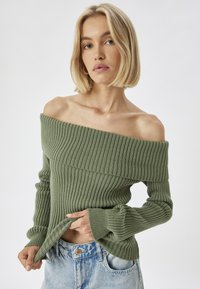 Olivenfarbener, schulterfreier, gerippter Pullover mit langen Ärmeln und weitem Halsausschnitt, kombiniert mit hellblauen, distressed Jeans.