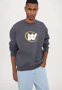 Grijze sweatshirt met een groot wit en oranje logo op de voorkant. Heeft een ronde halslijn en geribde manchetten. Gecombineerd met lichtblauwe jeans.