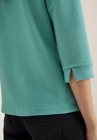 Top de tela acanalada en turquesa con mangas de tres cuartos y aberturas laterales, combinado con pantalones negros, que exhibe una textura suave.