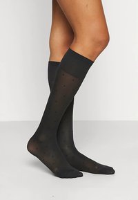 Svarte knelangssokker laget av gjennomsiktig materiale med polkaprikker. Strukturert ribbestrikket topp for bedre grep og komfort.