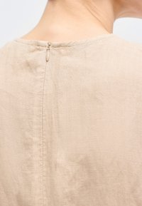 Πίσω γραμμή λαιμού μπεζ λινής ενδυμασίας με μικρό μπεζ φερμουάρ στο κέντρο της ραφής, φορεμένο από άτομο με ανοιχτό δέρμα.