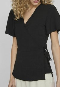 Top avvolgente nero realizzato in tessuto texturizzato, con maniche corte a sbuffo e dettaglio con fiocco laterale. Design a V con un sottile motivo stropicciato.
