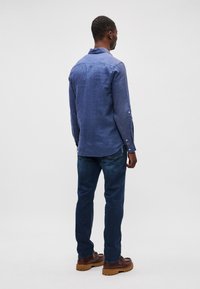 Μπλε λινό πουκάμισο με μακριά μανίκια, κλασικό γιακά και κουμπιά στους καρπούς, συνδυασμένο με σκούρες μπλε τζιν και καφέ δερμάτινες μπότες με κορδόνια.