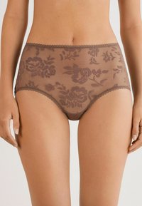 Bruine doorschijnende high-waist onderbroek met bloemmotief, gedragen door een staand persoon met ontspannen handen aan de zij.
