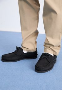 Mustad nahast loaferid ümmarguse varbaga, nööridega ja pehme tekstuuriga. Kombineeritud heledate pükstega, luues kontrasti sinise taustaga.