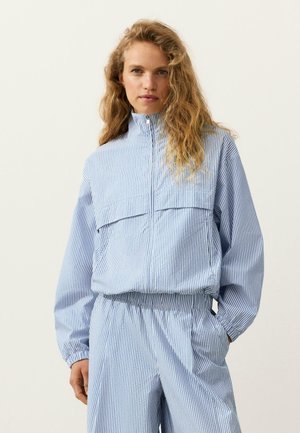 Vrouw met een lichtblauw-wit verticaal gestreept ritssluitingjack en bijpassende elastische taillebroek, staand met één hand in de zak.