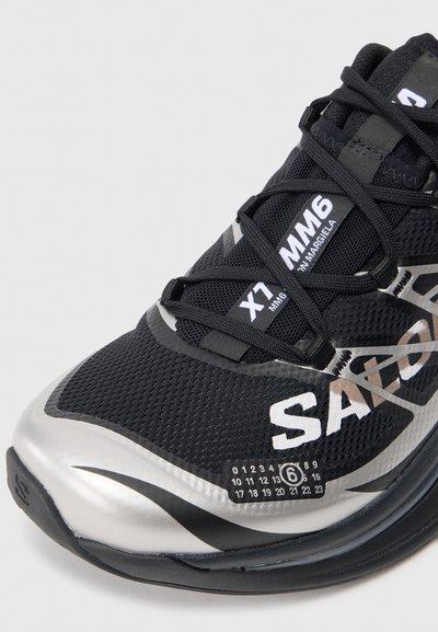 Černá sportovní bota s síťovaným svrškem, stříbrnými akcenty a texturovanou podrážkou. Obsahuje logo a detaily modelu na boku.