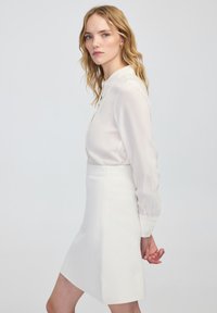 Weißer, taillierter Minirock kombiniert mit einer leichten, transparenten Bluse. Die Bluse hat einen klassischen Kragen und lange Ärmel mit Manschettendetails.