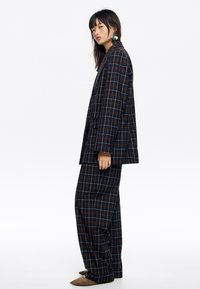 Granatowa marynarka w kratę i szerokie spodnie z pomarańczowymi akcentami, wykonane z miękkiej tkaniny. Dopasowany krój z wyraźnie zarysowaną formą i kontrastującymi teksturami.