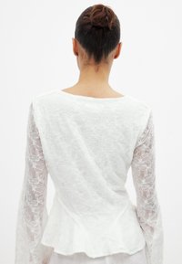 Weiße Bluse mit langen Ärmeln und floralem Spitzenmuster. Glatte Textur, Peplum-Saum, runder Ausschnitt und durchsichtige Ärmel. Rückansicht gezeigt.