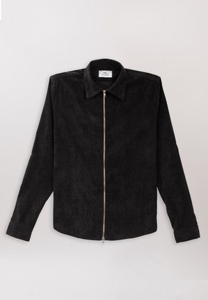 Svart skjorte med krage laget av myk, teksturert stoff med glidelås foran. Har lange ermer og rett kant.