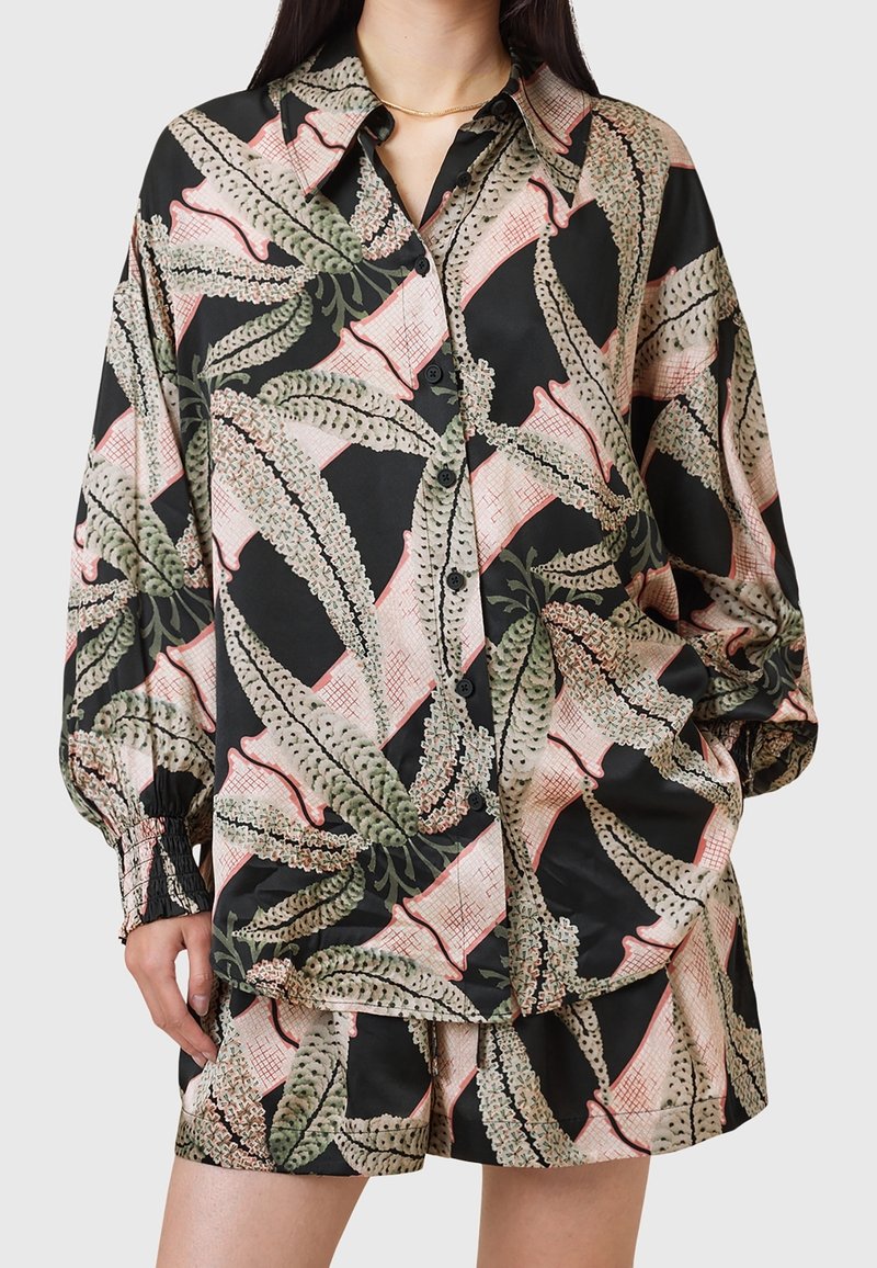 Femme portant un chemisier noir ample et un short avec un imprimé botanique abstrait rose et vert, posant avec une main dans la poche.