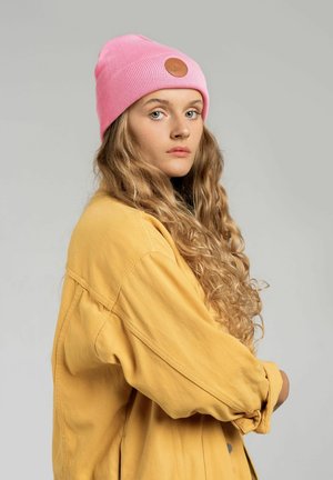 Różowy prążkowany beanie z skórzaną łatką, noszony przez osobę z długimi, falującymi włosami, w połączeniu z żółtą, oversize'ową koszulą. Neutralne tło.