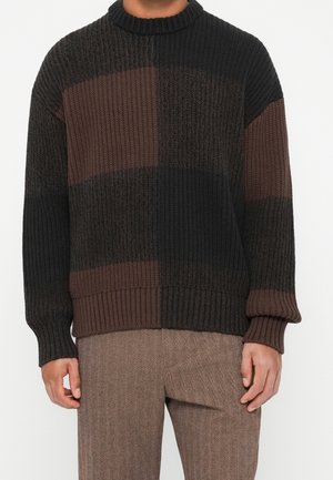 Homme portant un pull en maille épaisse avec des blocs de couleur noir et marron foncé et un pantalon marron texturé, debout devant un fond blanc.