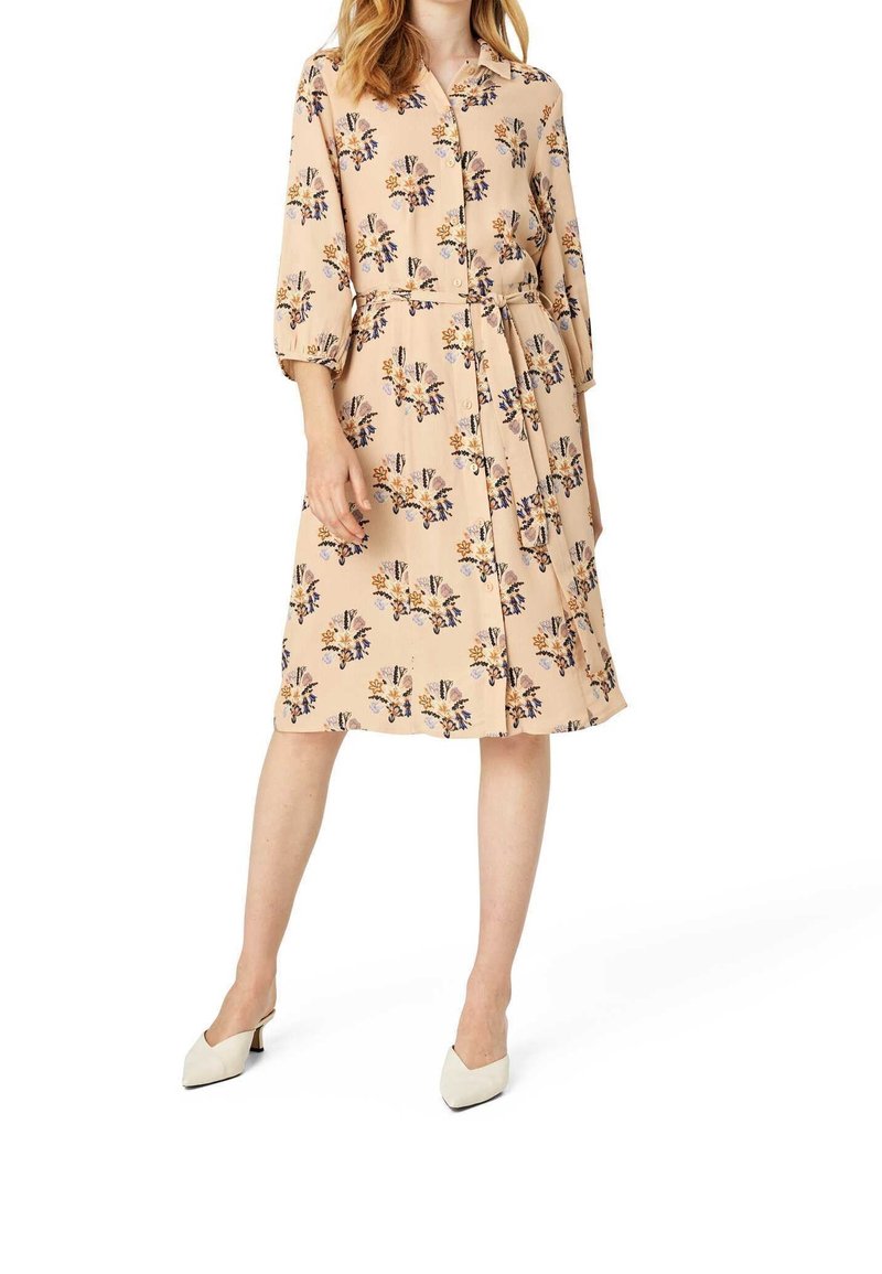Femme portant une robe beige fleurie boutonnée avec une ceinture à la taille et des manches trois-quarts, associée à des chaussures blanches à bout pointu.