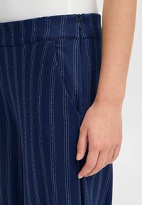 Pantalons taille haute rayés bleu marine, avec une texture lisse, présentant des fines rayures blanches, des poches latérales et une fermeture éclair à l'arrière.