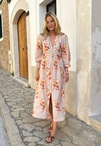 Florale midi-jurk in zacht roze met oranje en bordeaux bloemen, lange mouwen en knoopdetails aan de voorkant. Gepaard met bruine sandalen.