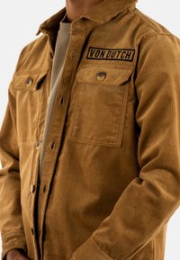 Chaqueta de pana marrón con cuello, dos bolsillos frontales y un parche del logo "VON DUTCH" en negro. Textura suave y patrón acanalado.