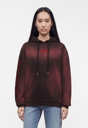 Femme aux longs cheveux foncés portant un sweat à capuche dégradé rouge foncé avec poche avant et motifs géométriques rouges sur la manche gauche, assorti à un jean bleu clair.
