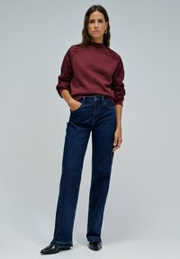 Mujer vestida con un suéter de punto burdeos con bordados florales en las mangas y jeans rectos de color azul oscuro, de pie con una mano en el bolsillo.