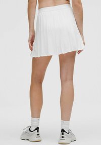 Jupe plissée blanche avec une taille élastique, longueur genou, texture lisse. Portée avec des baskets grises et des chaussettes blanches.